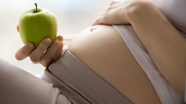Ernaehrung-Schwangerschaft_Web800-800×400.jpg Eine schwangere Person hält einen grünen Apfel und berührt sanft ihren Bauch.