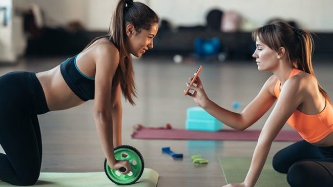 Sporthygiene_Web800-800×400.jpg Zwei Frauen trainieren in einem Fitnessstudio, eine benutzt ein grünes Trainingsgerät und die andere filmt mit ihrem Handy.
