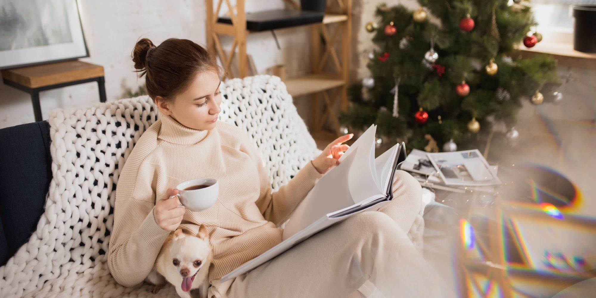Immer_mit_der_Ruhe.jpg Eine Frau in bequemer Kleidung sitzt auf einem Sofa, liest in einem Buch und hält eine Tasse Kaffee, während ein Hund bei ihr sitzt.