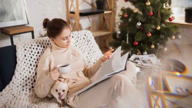 Immer_mit_der_Ruhe.jpg Eine Frau in bequemer Kleidung sitzt auf einem Sofa, liest in einem Buch und hält eine Tasse Kaffee, während ein Hund bei ihr sitzt.