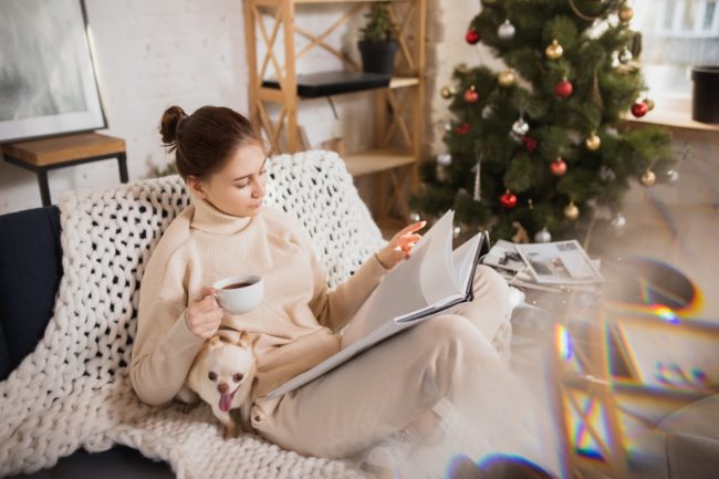 Immer_mit_der_Ruhe.jpg Eine Frau in bequemer Kleidung sitzt auf einem Sofa, liest in einem Buch und hält eine Tasse Kaffee, während ein Hund bei ihr sitzt.