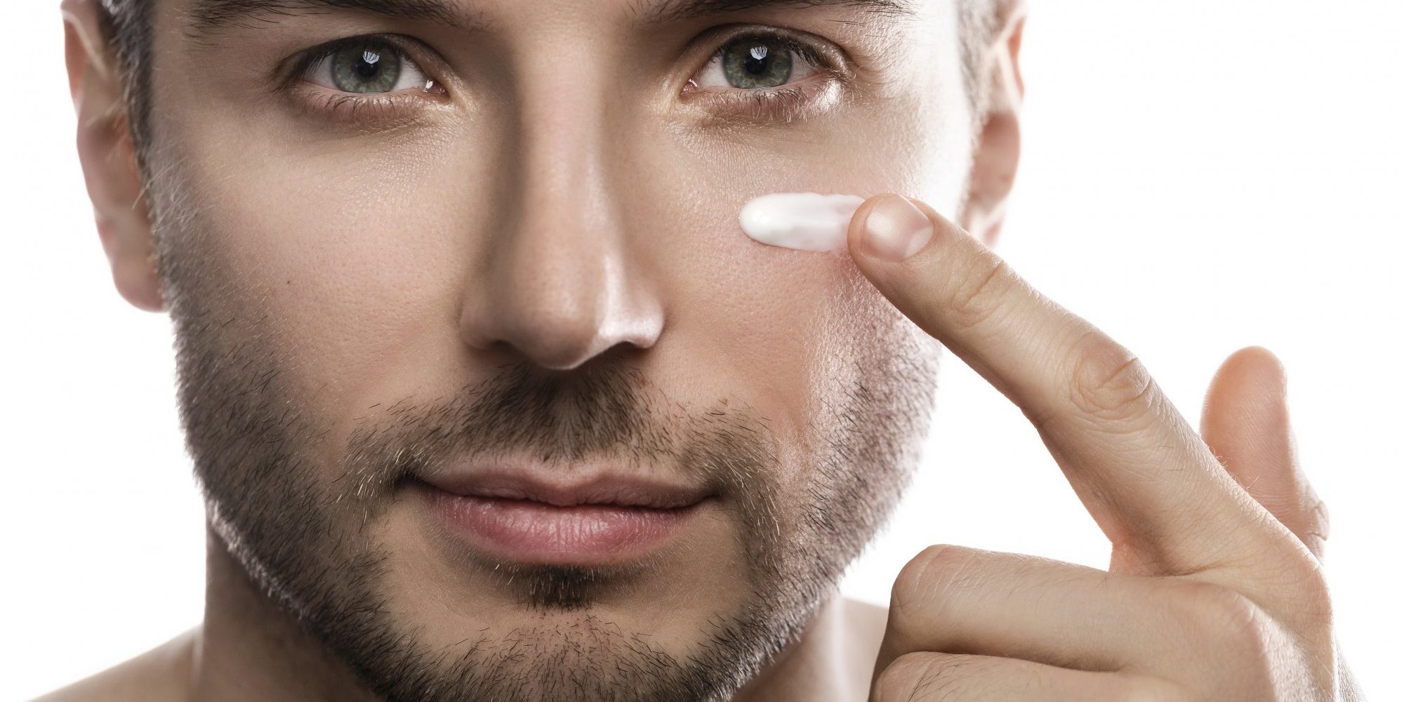 Ein Mann trägt eine Creme auf seine Wange auf, während er in die Kamera schaut. Hintergrund ist neutral.