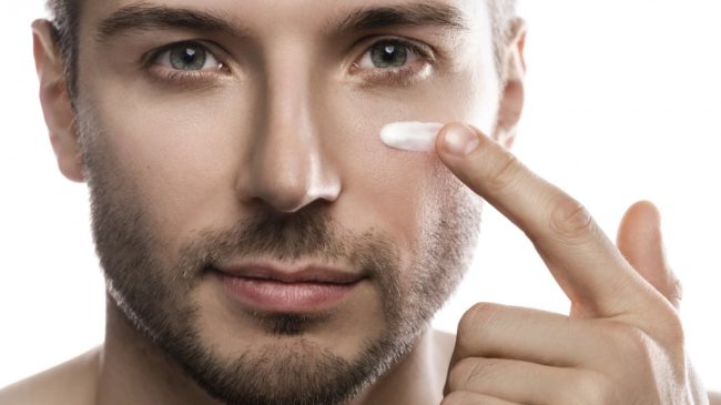 Verletzliche_Maennerhaut.jpg Ein Mann trägt eine Creme auf seine Wange auf, während er in die Kamera schaut. Hintergrund ist neutral.