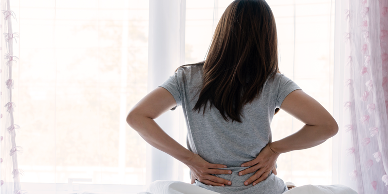 Eine Person mit langen Haaren sitzt auf einem Bett und hält sich den Rücken vor einem Fenster.
