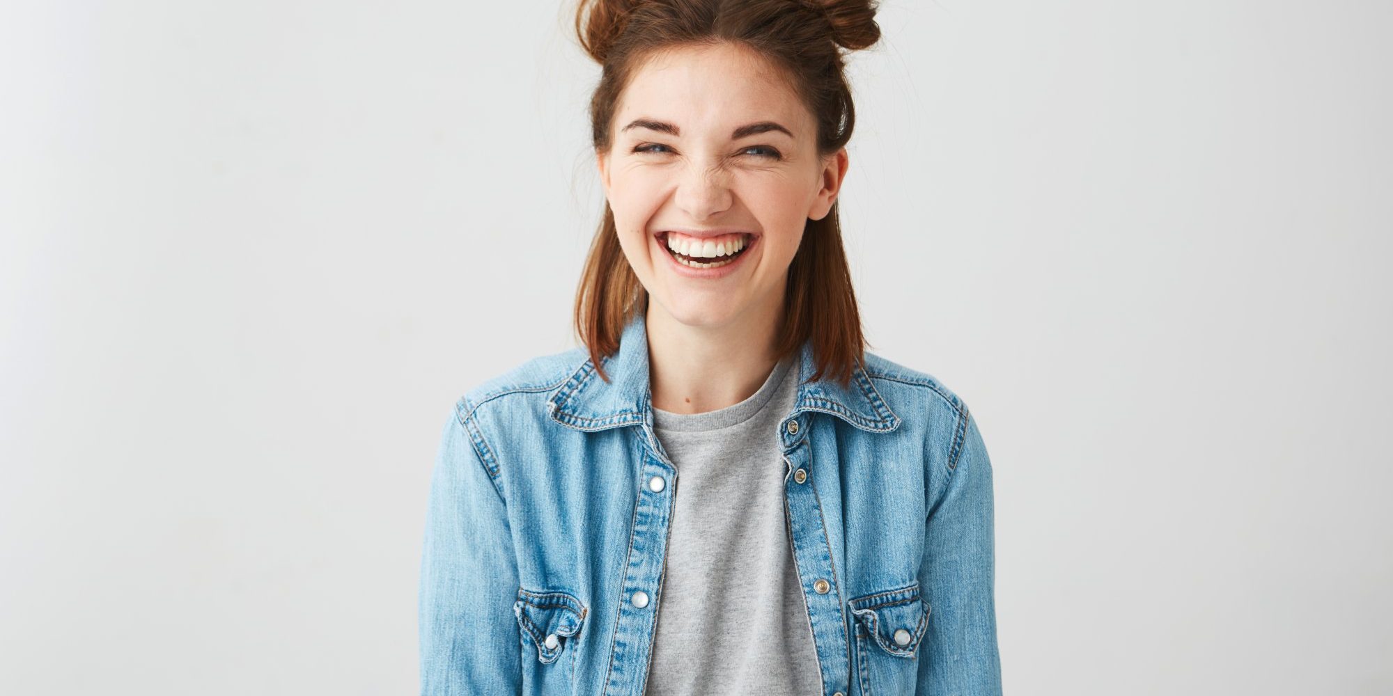Lachen.jpg Lachende junge Frau mit Duttfrisur, trägt eine Jeansjacke über einem grauen T-Shirt, vor einem hellen Hintergrund.