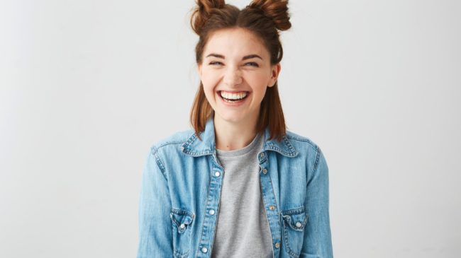 Lachen.jpg Lachende junge Frau mit Duttfrisur, trägt eine Jeansjacke über einem grauen T-Shirt, vor einem hellen Hintergrund.