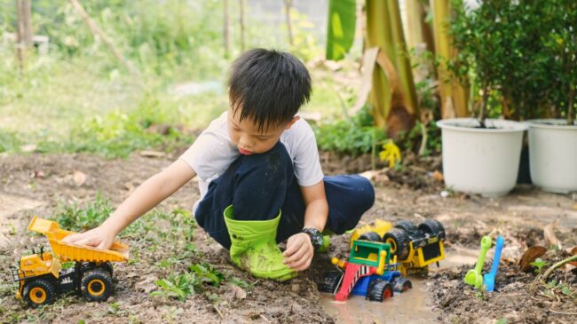 wie-viel-dreck-ist-gesund.jpg Ein Junge im Garten spielt mit verschiedenen Spielzeugfahrzeugen, darunter ein Lkw und ein Bagger, während er in matschigem Boden sitzt.