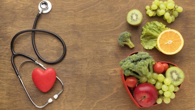 Ein Stethoskop und ein rotes Herzmodell neben einer herzförmigen Schale mit frischen Obst- und Gemüsesorten.