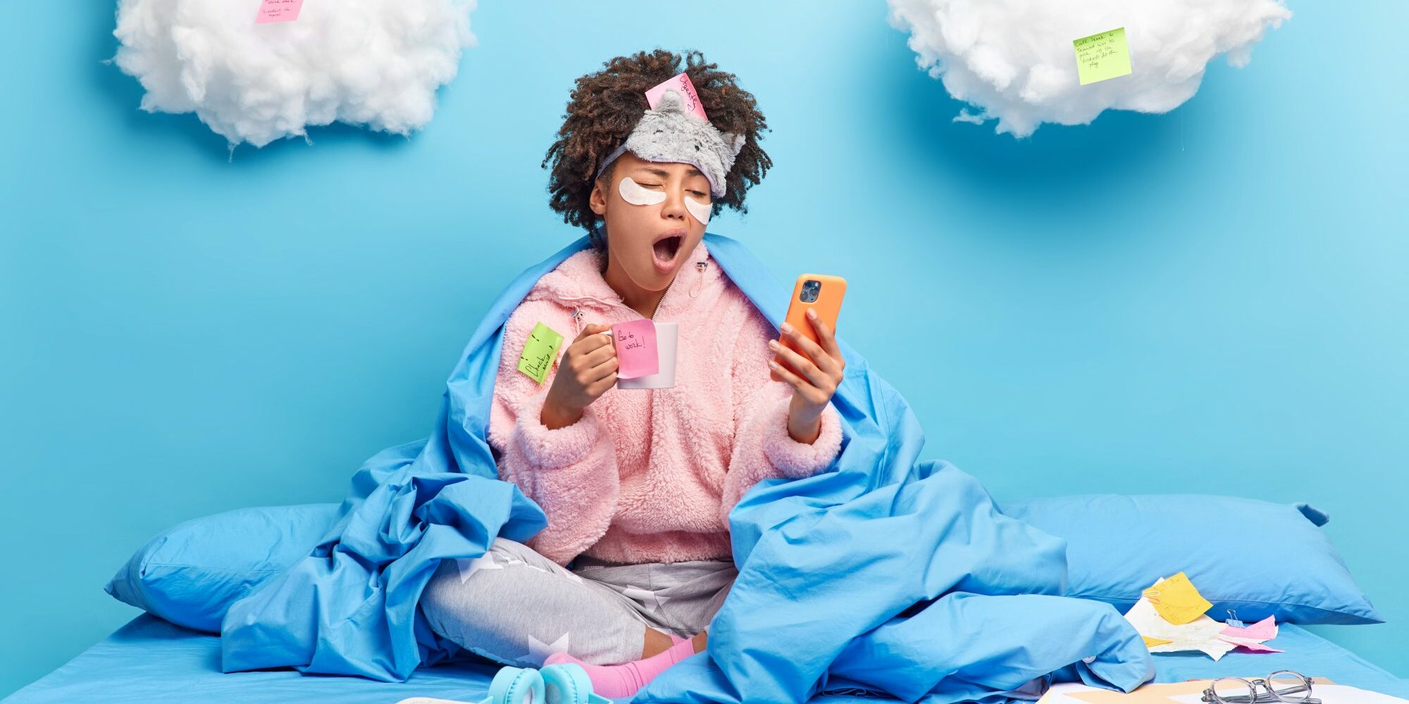 Eine Person in einem blauen Bett, umhüllt von einer Decke, schaut müde auf ihr Handy und hält eine Tasse in der Hand. Wolken mit Haftnotizen im Hintergrund.