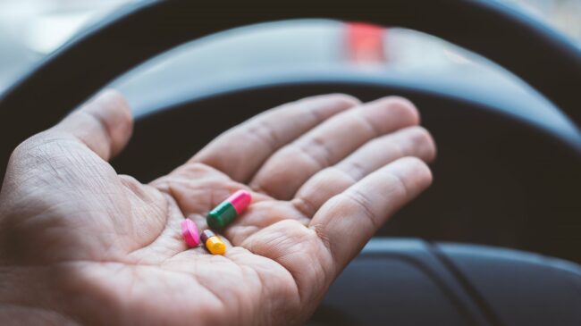 Arzneimittel_im_Strassenverkehr.jpg Eine Hand hält bunte Kapseln über dem Lenkrad eines Fahrzeugs.