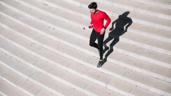 Stiegensteigen_Fitness.jpg Mann in rotem Shirt joggt über eine helle Treppe, sein Schatten ist deutlich sichtbar.