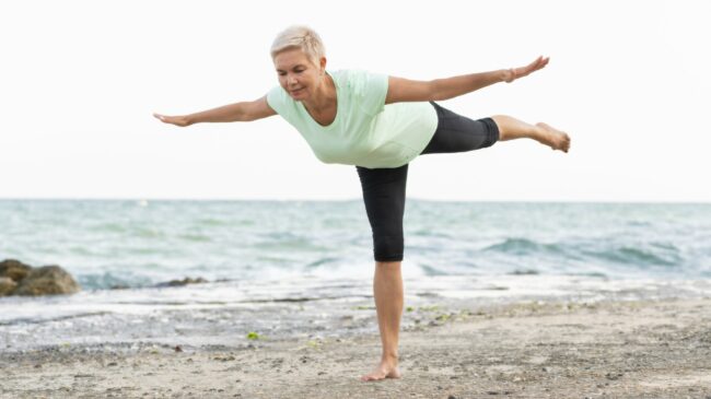 Balancetraining2.jpg Eine Frau in einem hellgrünen T-Shirt und schwarzen Leggings balanciert auf einem Bein am Strand.