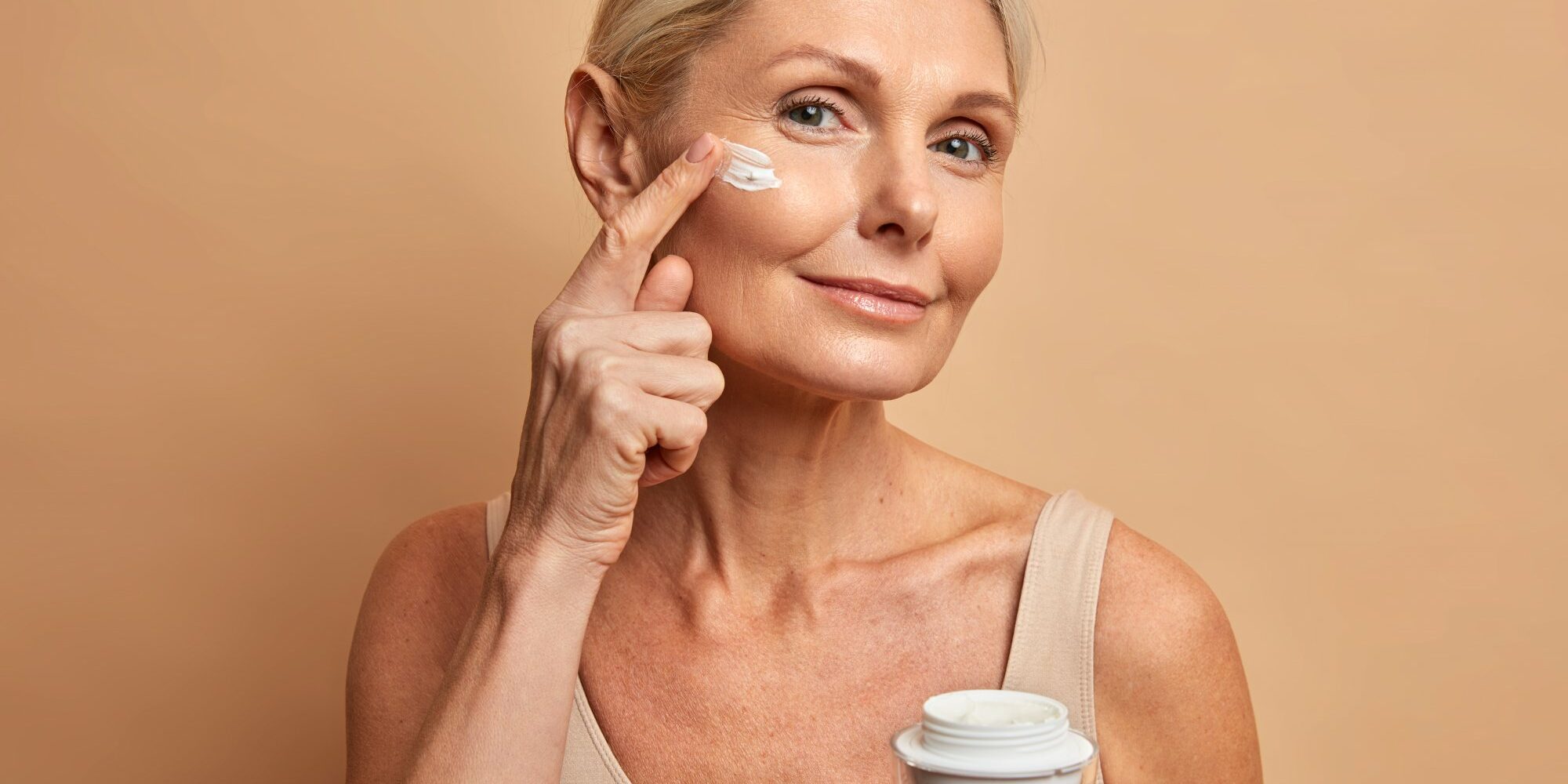 Anti-Aging.jpg Eine Frau mit blondem Haar trägt Gesichtspflegecreme auf ihre Wange auf, während sie einen Cremetiegel in der Hand hält.