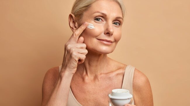 Anti-Aging.jpg Eine Frau mit blondem Haar trägt Gesichtspflegecreme auf ihre Wange auf, während sie einen Cremetiegel in der Hand hält.
