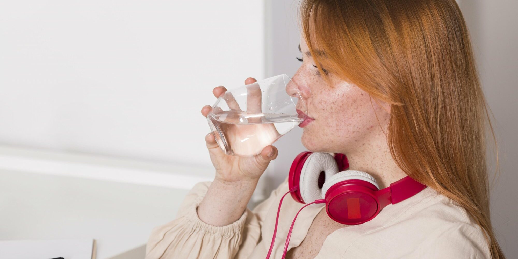 Eine Person mit roten Kopfhörern trinkt aus einem Glas Wasser und sitzt an einem Schreibtisch.