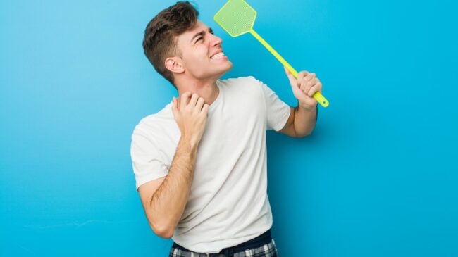 plagegeister.jpg Junger Mann in einem weißen T-Shirt hält ein grünes Insektenspray mit einem lächelnden Gesicht vor einer blauen Wand.