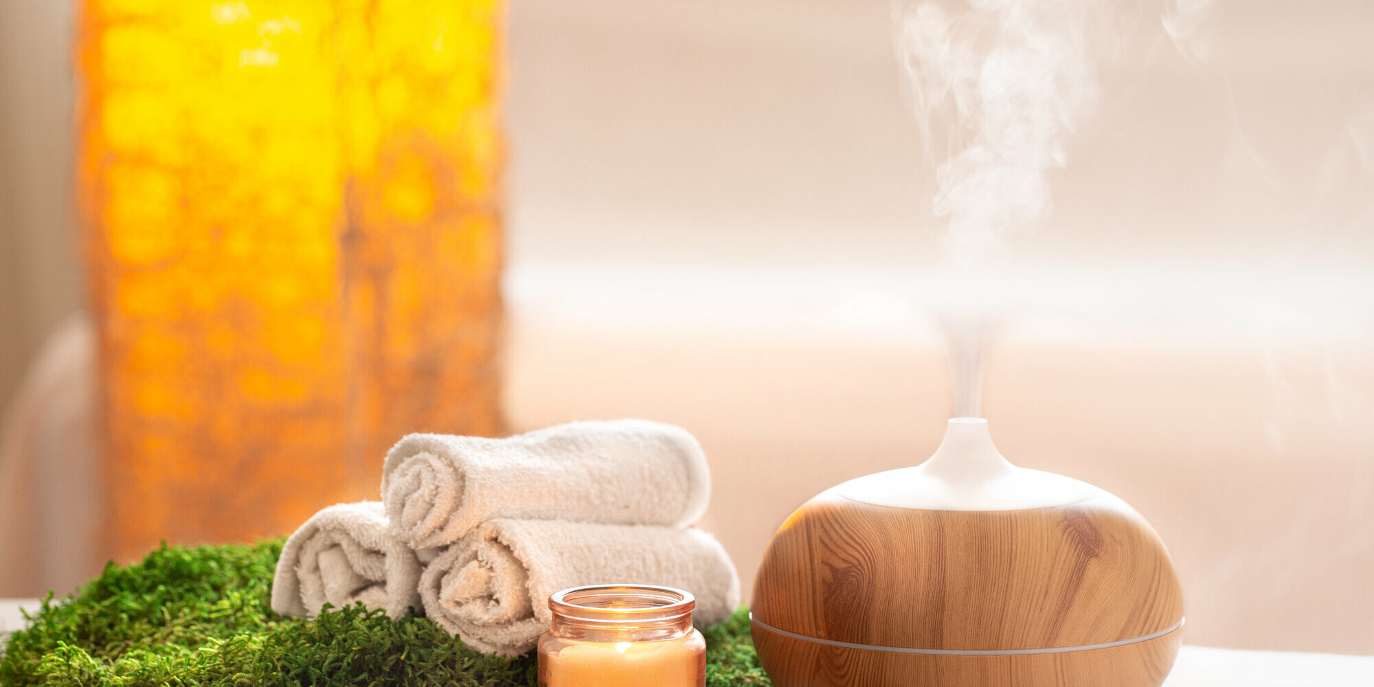 aromatherapie.jpg Ein Aromadiffusor aus Holz mit aufsteigendem Dampf, umgeben von Handtüchern, einer Kerze und grünem Moos.