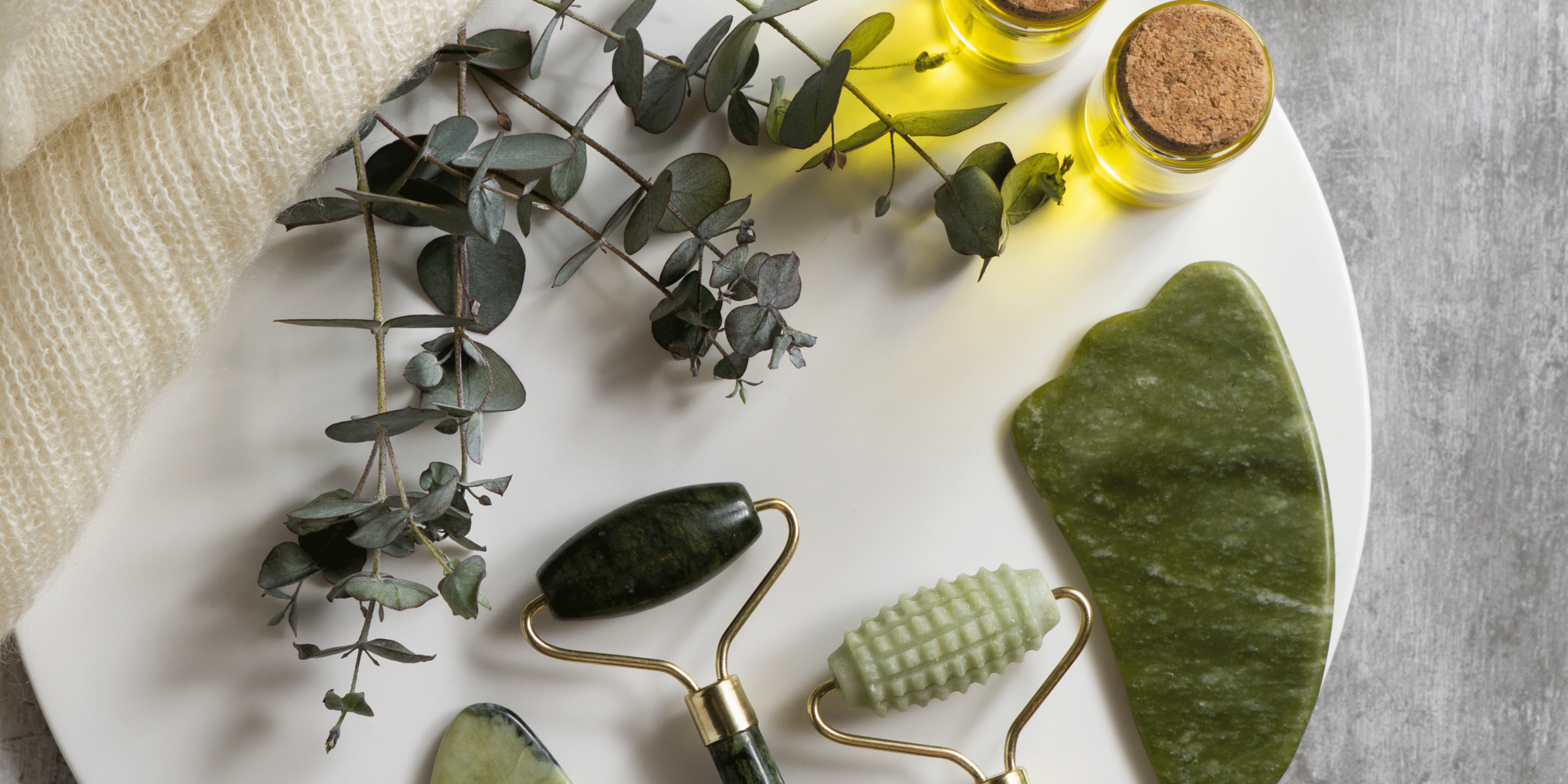 nachhaltige-kosmetik.png Ein weißer Teller mit drei Jade-Rollern, einem gua sha Stein, zwei Fläschchen ätherischem Öl und Eukalyptuszweigen.