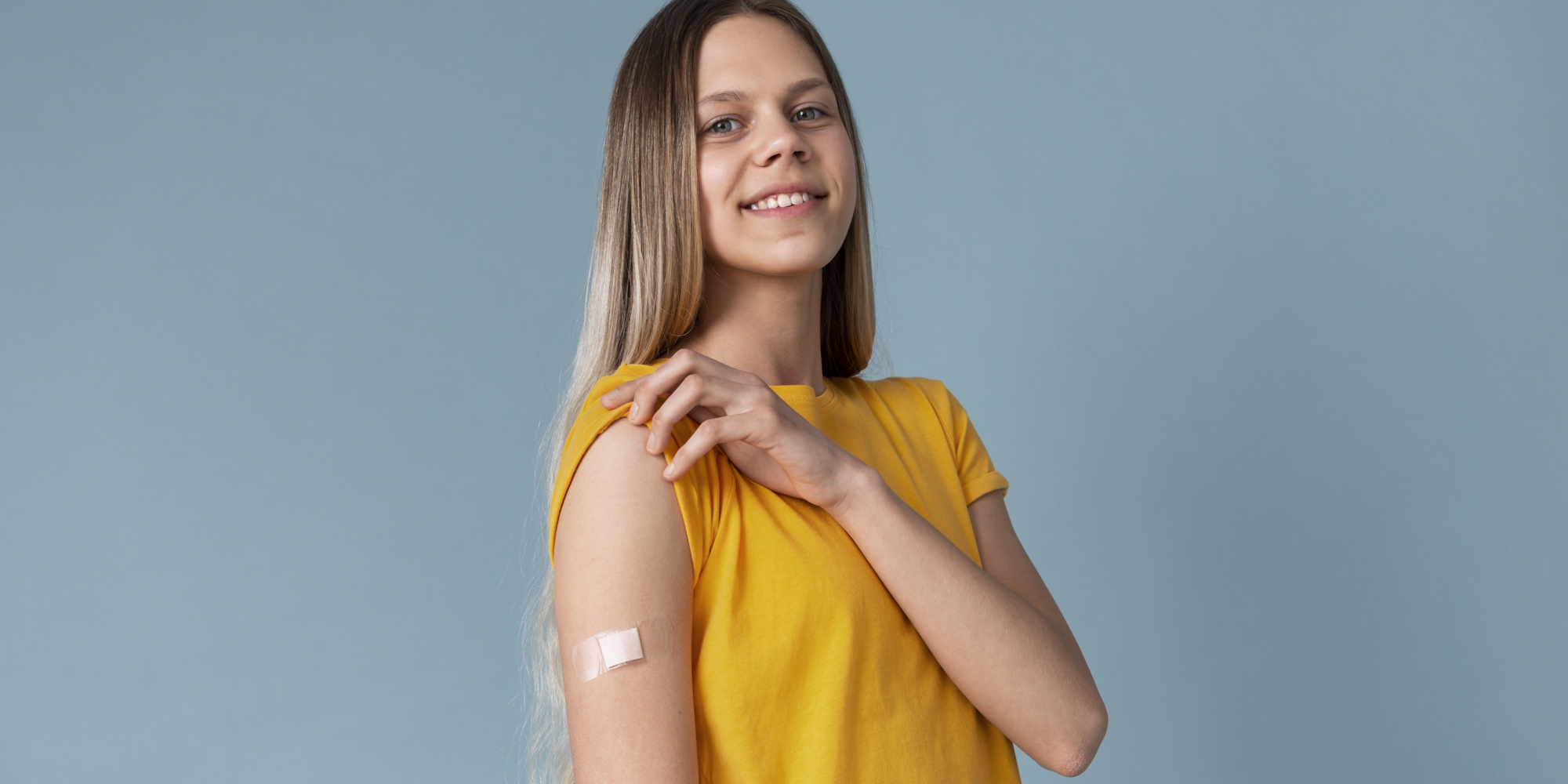HPV_Impfung.png Eine lächelnde junge Frau mit langen Haaren trägt ein gelbes T-Shirt und zeigt einen Verband am Oberarm.