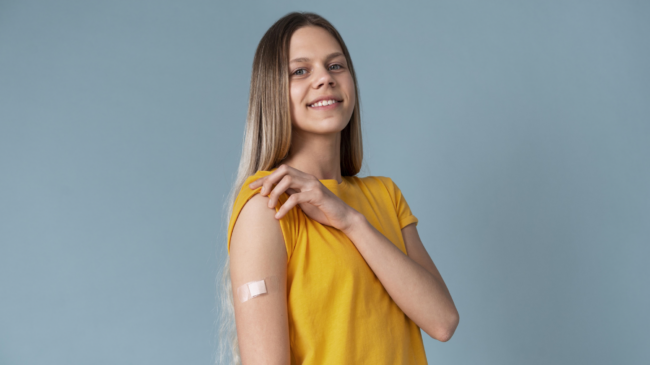 Eine lächelnde junge Frau mit langen Haaren trägt ein gelbes T-Shirt und zeigt einen Verband am Oberarm.