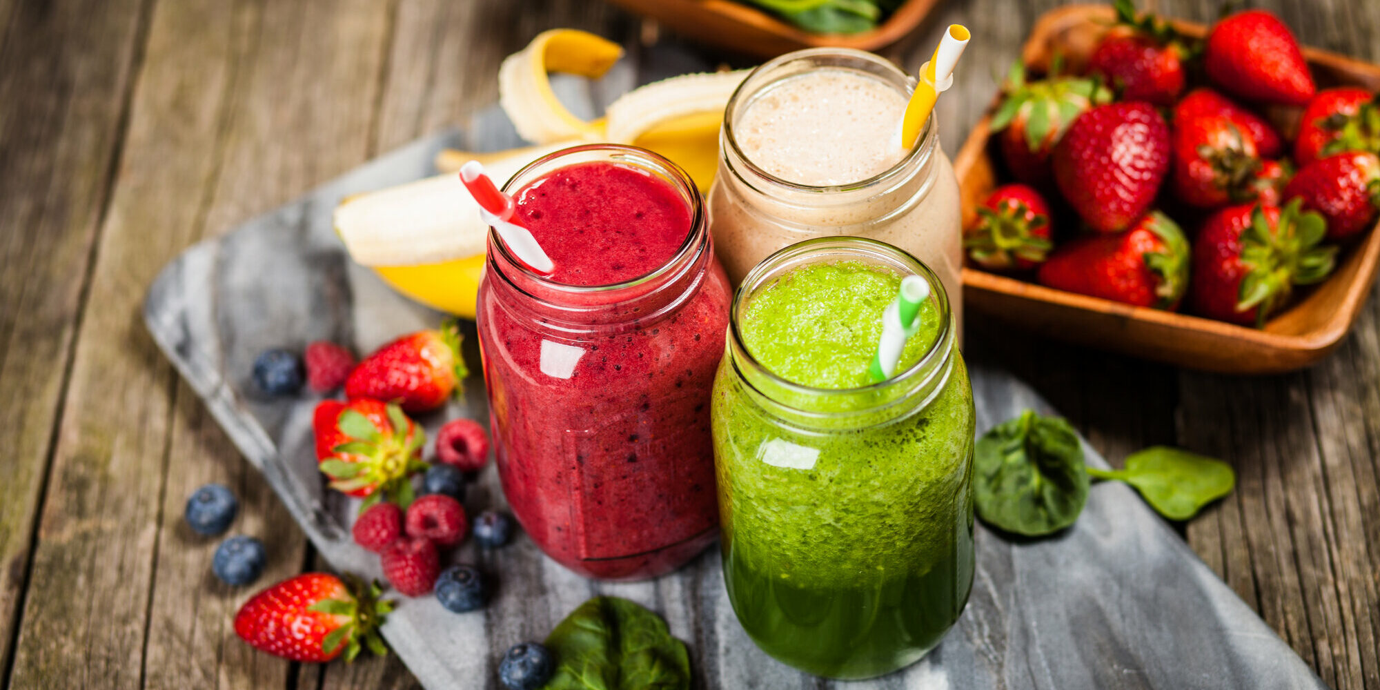 Smoothies__2_.jpeg Drei bunte Smoothies in Gläsern mit Strohhalmen, umgeben von frischen Früchten und Spinat auf einer Holzunterlage.