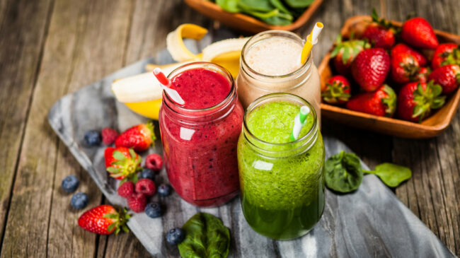 Drei bunte Smoothies in Gläsern mit Strohhalmen, umgeben von frischen Früchten und Spinat auf einer Holzunterlage.