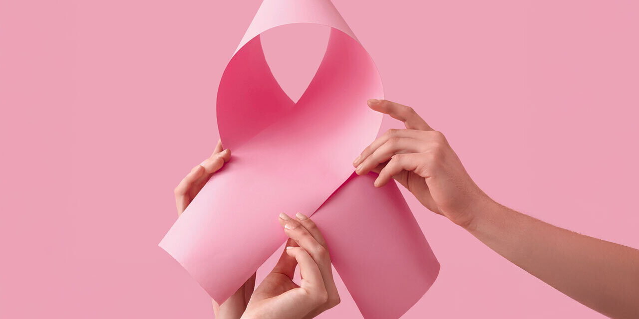 Welt-Brustkrebs-Tag.jpg Zwei Hände halten ein rosa Papier, das zu einer Schleife geformt ist, vor einem pinken Hintergrund.