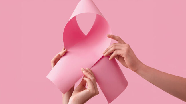 Zwei Hände halten ein rosa Papier, das zu einer Schleife geformt ist, vor einem pinken Hintergrund.