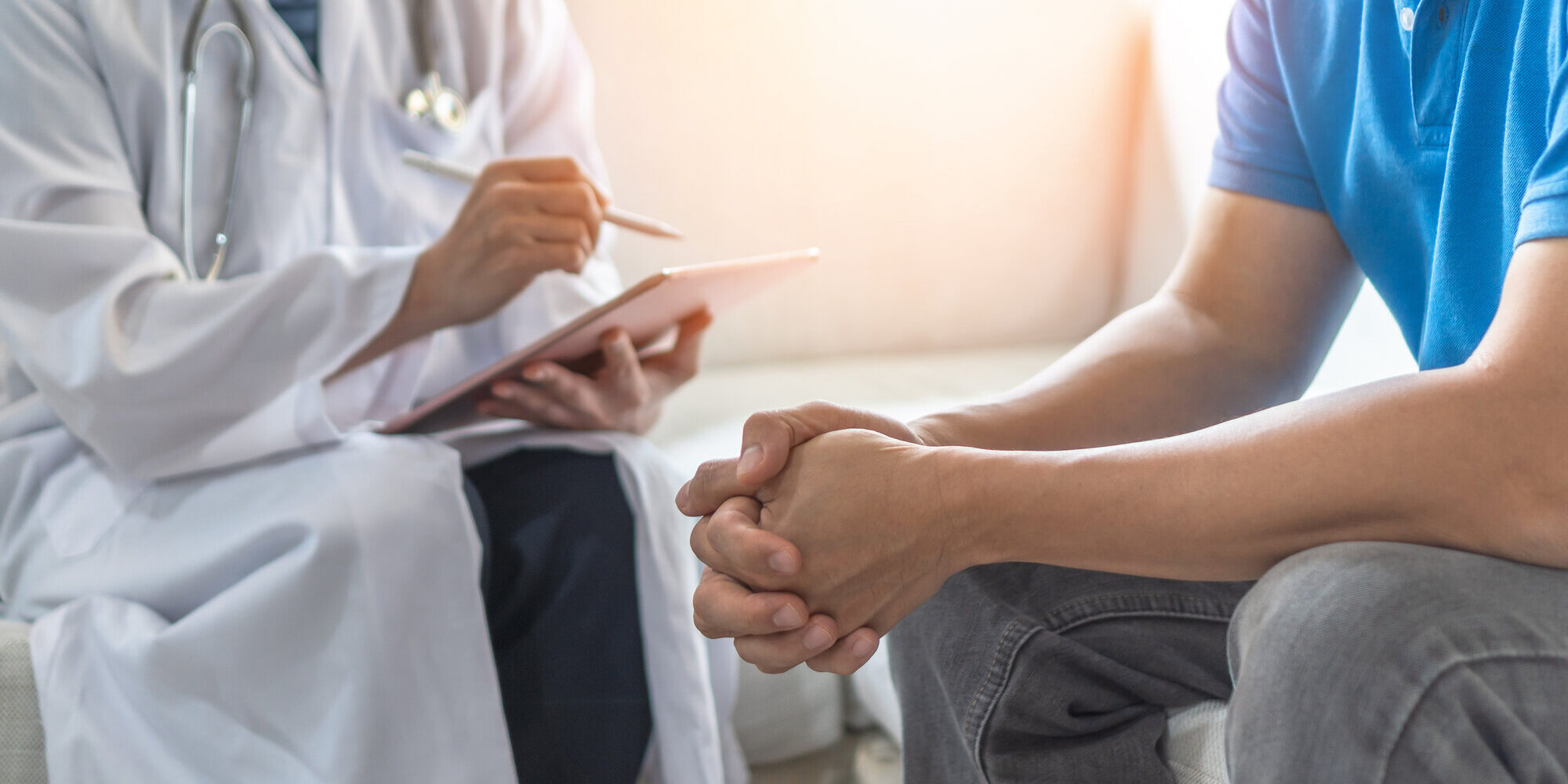 maenner-gesundheit.jpeg Arzt in weißem Kittel notiert auf einem Tablet, während ein Patient mit zusammengefalteten Händen sitzt.