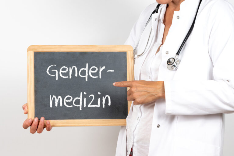Eine Person in Arztkleidung hält eine Tafel mit der Aufschrift "Gendermedizin".