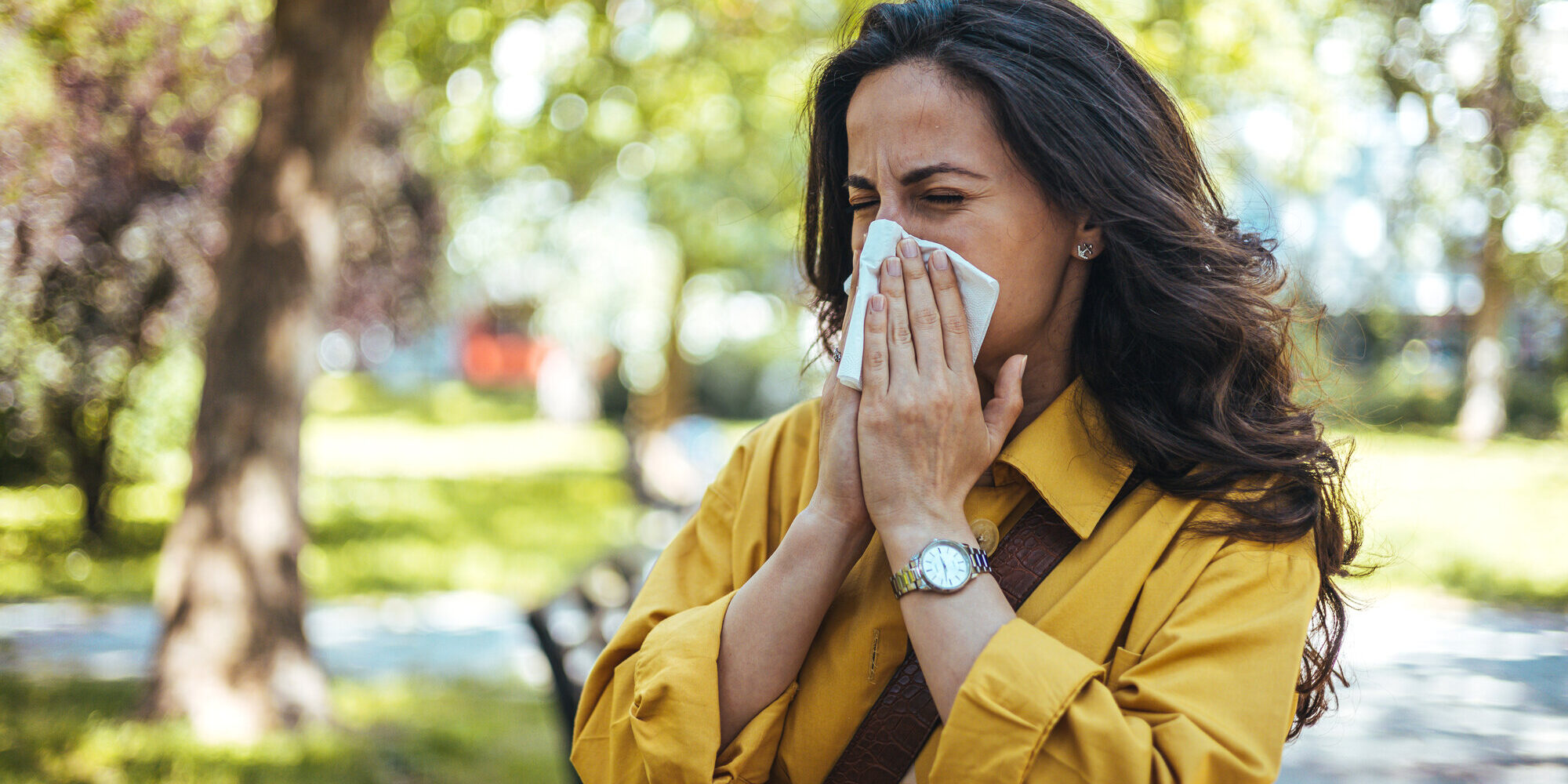 pollenallergie.jpeg Frau in einem gelben Hemd, die sich mit einem Taschentuch die Nase putzt, umgeben von grüner Parklandschaft.