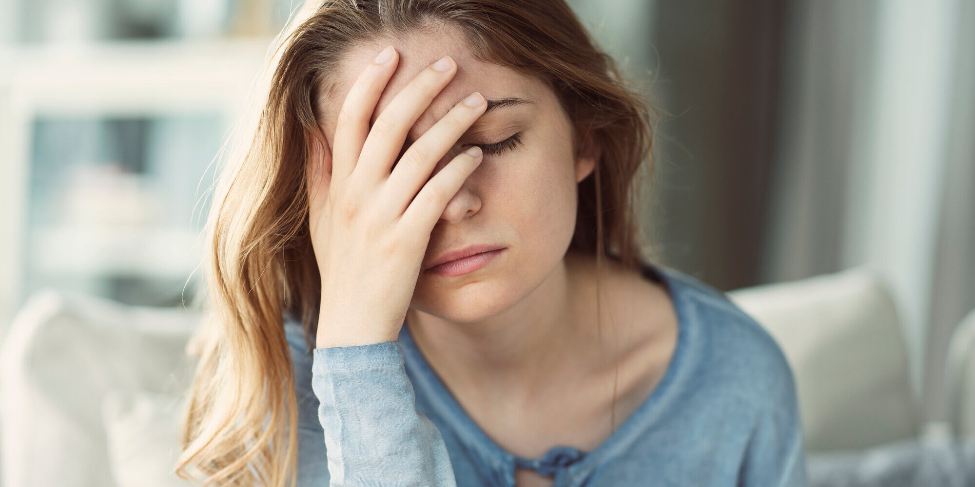 Junge Frau mit langen Haaren und blauem Oberteil, die frustriert ihre Hand an die Stirn legt, im Innenraum sitzend.